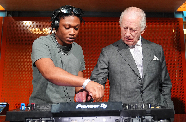 King Charles trying his hand at a DJ turntable, standing next to a professional DJ at a royal charity music event.